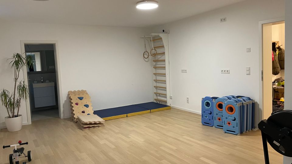 Heller, leerer Innenraum mit hellem Holzboden und weißen Wänden, ausgestattet wie ein Spiel- oder Bewegungsraum mit Sprossenwand, Turnringen und Turnmatten. Links und rechts führen Türöffnungen in angrenzende Räume; an der Wand stehen zusammengeklappte Spielgitter-Elemente und ein Pflanzenkübel.