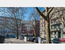 Straßenszene mit mehrstöckigen Wohnhäusern im Hintergrund, teils in weißem Putz und rotem Akzent, sowie einem Fernsehturm, der hinter den Bäumen sichtbar ist. Im Vordergrund stehen auf einem Parkplatz mehrere Autos und kahle Bäume werfen Schatten auf das Straßenpflaster.