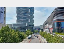 Straßenszene in einem modernen Stadtviertel mit einem hohen Bürogebäude aus Glas und Stahl in der Mitte, flankiert von einem futuristischen Gebäude mit geschwungenen, metallischen Fassaden rechts. Im Vordergrund befinden sich Bäume und eine Straße mit parkenden Autos.