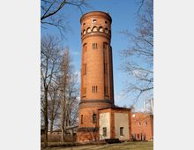 Roter Backsteinturm mit Rundbogenfenstern und dekorativen Ziegelmustern, vermutlich ein ehemaliger Wasserturm, umgeben von kahlen Bäumen und angrenzenden flachen Gebäuden. Der Himmel ist klar und blau.