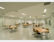 Ein großer, heller Speisesaal mit schlichtem Design, ausgestattet mit Holztischen und passenden Holzstühlen auf einem hellen Boden. Die Decke ist mit integrierten Beleuchtungselementen und Belüftungsgittern versehen, und der Raum wird durch weiße Pfeiler unterteilt.