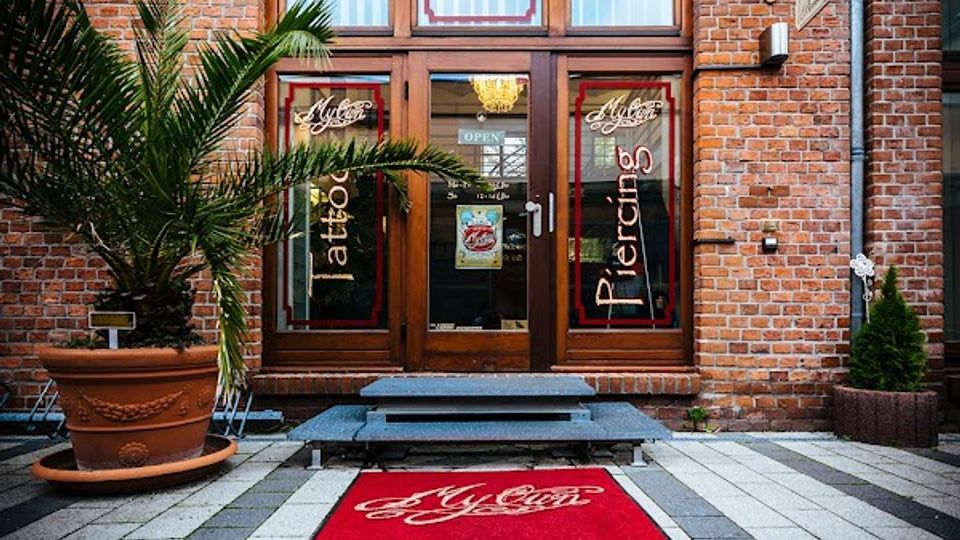 Eingang eines Backsteingebäudes mit zweiflügeliger Glastür und großen Fenstern darüber, versehen mit Schriftzügen für Tattoo- und Piercing-Studio. Davor liegt ein roter Teppich auf einem gepflasterten Hof; links steht eine große Pflanze im Kübel.