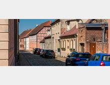 Blick entlang einer schmalen Kopfsteinpflasterstraße in einem Altstadtviertel, gesäumt von dicht stehenden Wohnhäusern mit Putzfassaden und teils sichtbarem Fachwerk sowie Ziegelmauern. Am Straßenrand parken mehrere Autos; Laternen und rot gedeckte Satteldächer prägen das Straßenbild.