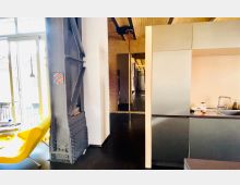 Moderne Wohnung mit industriellen Elementen. Sichtbare Stahlträger, eine gelbe Sitzgelegenheit vor einer großen Fensterfront, eine offene Küche mit grauer Oberflächenverkleidung und Wände aus Backstein und hellem Holz prägen den Raum.