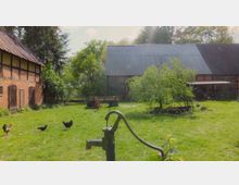 Ein ländlicher Hof mit zwei Fachwerkgebäuden, dazwischen eine Grünfläche mit einem kleinen Baum, einer alten Wasserpumpe im Vordergrund sowie Hühnern und einem Hahn auf der Wiese. Im Hintergrund sind Bäume und ein Anhänger sichtbar.