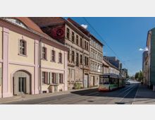 Straßenszene in einer Stadt mit einer gelb-rosa Fassade eines renovierten Gebäudes und einem angrenzenden verfallenen Ziegelgebäude. Eine Straßenbahn fährt auf Gleisen entlang der Straße, während im Hintergrund weitere Gebäude und ein klarer blauer Himmel zu sehen sind.