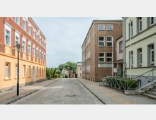 Straßenszene mit gepflasterter Fahrbahn und Schulgebäuden auf beiden Seiten, darunter ein Backsteinbau und ein beige verputztes Gebäude mit der Aufschrift „Oberschule Theodor Fontane“. Im Hintergrund sind Bäume und ein kleiner, älterer Anbau erkennbar.