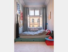 Ein helles Schlafzimmer mit einem niedrigen Bett auf einer türkisfarbenen Plattform, gestreifter Bettwäsche und einem Fenster mit Blick auf ein Backsteingebäude. Die Wände sind mit Bildern dekoriert, und Vorhänge rahmen das Fenster ein; auf dem Boden liegt ein heller Teppich.