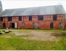 Backstein-Gebäude mit Fachwerk-Elementen, mehreren Holztoren und kleinen Fenstern in ländlicher Umgebung. Im Vordergrund befindet sich eine grasbewachsene Fläche mit Baumstümpfen und vereinzelten Gegenständen.