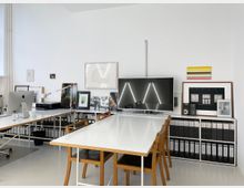 Modernes Büro mit weißen Wänden und einem minimalistischen Design. Ein großer Tisch mit Holzkanten und Stühlen steht im Vordergrund, im Hintergrund befinden sich ein Regal mit Aktenordnern, ein Flachbildschirm sowie verschiedene gerahmte Bilder und Kunstwerke.