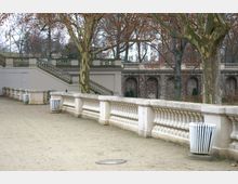 Ein parkähnlicher Bereich mit einer breiten, gepflasterten Wegfläche, begrenzt durch weiße Steinbalustraden. Dahinter befinden sich eine Treppe, mehrere steinerne Bögen und große Bäume mit herbstlichem Laub.