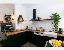 Moderne Küche mit dunklen Schränken und Arbeitsplatten aus Holz, einer weißen Keramikspüle und einer weißen Fliesenrückwand. Dekorative Elemente wie Hängepflanzen, offene Regale mit Büchern und Gewürzen sowie eine Kaffeemaschine sind sichtbar.