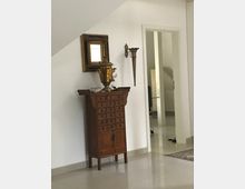 Innenansicht eines hellen Flurs mit glänzendem Fliesenboden und schräger Decke. An der Wand steht ein hohes Holzschränkchen mit vielen kleinen Schubladen, darüber ein gerahmter Spiegel und eine Wandlampe; rechts führt eine offene Tür in einen weiteren Raum.