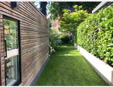 Holzverkleidetes Gebäude mit Glasfenster, das Flaschen in einem Kühlschrank zeigt, auf der linken Seite. Rechts davon ein gepflegter, schmaler Rasenstreifen, begrenzt durch eine Hecke und eine niedrige Mauer. Im Hintergrund sind Bäume und ein Teil eines Hauses zu sehen.