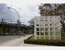 Weiße kubische Skulptur aus Gitterelementen auf einer Grünfläche, daneben eine Straße mit Autos und Gehweg. Im Hintergrund eine erhöhte Brücke mit einem Flugzeug sichtbar sowie Gebäude und Bäume.