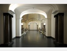 Flur im Innenbereich eines historischen Gebäudes mit gewölbter Decke, verzierten Säulen und dekorativen Geländern aus Metall. Der Gang führt zu einem Bogen, der sich in eine helle, stilisierte Wandöffnung öffnet.