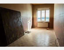 Leerer, renovierungsbedürftiger Innenraum mit gemusterten Tapeten, Parkettboden und einem großen Fenster über einem Heizkörper. An der linken Wand lehnt eine große dunkle Platte, daneben sind beschädigte Wandstellen und Bauschutt sichtbar.