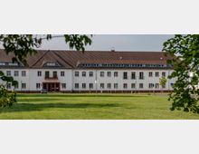 Ein zweistöckiges, symmetrisches Gebäude mit weißer Fassade und rotem Ziegeldach, umgeben von einer großen, offenen Grünfläche. Im Vordergrund befinden sich Bäume und im Hintergrund Straßenlampen sowie Parkschilder.