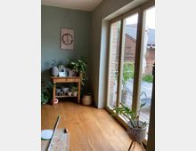 Ein gemütlicher Innenraum mit Holzboden, dekoriert mit Pflanzen auf einem kleinen Holztisch und in Körben. Große Glastüren führen zu einer Terrasse, auf der Ziegelwände und ein Teil eines Gebäudedachs sichtbar sind.