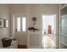 Helles Interieur eines Wohnflurs mit zwei Türen an gegenüberliegenden Wänden; links eine Doppeltür mit mattem Glaseinsatz, rechts eine Türöffnung mit Blick in ein Zimmer mit Holzboden, einem dunklen Holzschrank und einem Teppich. In der Mitte steht eine weiße Konsole, darüber eine Wanduhr in Form eines Kuckucksuhres.