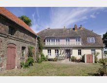 Ein Hof mit zwei Gebäuden: rechts ein zweistöckiges Wohnhaus mit heller Fassade, weißen Fensterläden und einem Balkon; links ein aus Naturstein gebautes Gebäude mit rotbrauner Tür und Ziegeldach. Im Vordergrund ein gepflasterter Hof mit Pflanzen und Sitzgelegenheiten.