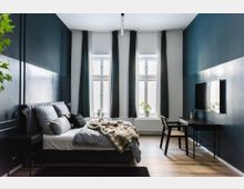 Modernes Schlafzimmer mit dunkelblauen Wänden, einem Doppelbett mit grauer Bettwäsche und einem schwarzen Nachttisch. Gegenüber dem Bett befindet sich ein Schreibtisch mit Stuhl sowie ein Flachbildfernseher, neben drei großen Fenstern mit langen Vorhängen und einem Zimmerpflanzentopf auf der Fensterbank.