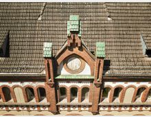 Backsteinfassade eines historischen Gebäudes mit Rundfenster und Zierelementen, darunter Arkadenfenster mit weißen Verzierungen. Das Dach ist mit dunklen Ziegeln und grünen Akzentfliesen an den Firsten gedeckt.