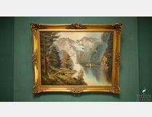 Ein Gemälde in einem goldenen, reich verzierten Rahmen zeigt eine idyllische Landschaft mit Bergen, einem See und einer kleinen Kapelle am Ufer. Der Hintergrund ist in einem matten Grünton gestrichen.