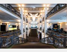 Großzügiges, zweistöckiges Atrium mit symmetrischen weißen Säulen, kunstvollen schwarzen Metallgeländern und dekorativen Kronleuchtern an der Decke. Zentral führt eine breite Treppe nach unten, während seitlich Sitzbereiche mit Sesseln und ein beleuchteter Empfangstresen erkennbar sind.