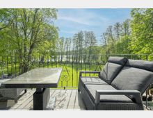 Blick von einer Terrasse mit schmiedeeisernem Geländer auf eine grüne Wiese, umgeben von hohen Bäumen. Im Vordergrund stehen Gartenmöbel aus Rattan mit grauen Polstern und ein Glastisch.