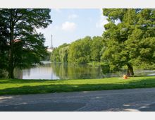 Ein Park mit einem großen Teich, umgeben von Bäumen, darunter Weiden, die über das Wasser hängen. Im Vordergrund ein gepflasterter Weg, eine Wiese mit einer Person, die auf einer Decke unter einem Baum sitzt, im Hintergrund ein hoher Turm sichtbar.