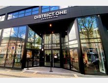 Moderne Fassade eines Coworking-Spaces mit der Aufschrift „District One Coworking“. Das Gebäude hat große Glasfenster, die die umliegenden Gebäude und Bäume spiegeln, sowie einen zentralen Eingangsbereich mit doppelten Glastüren.