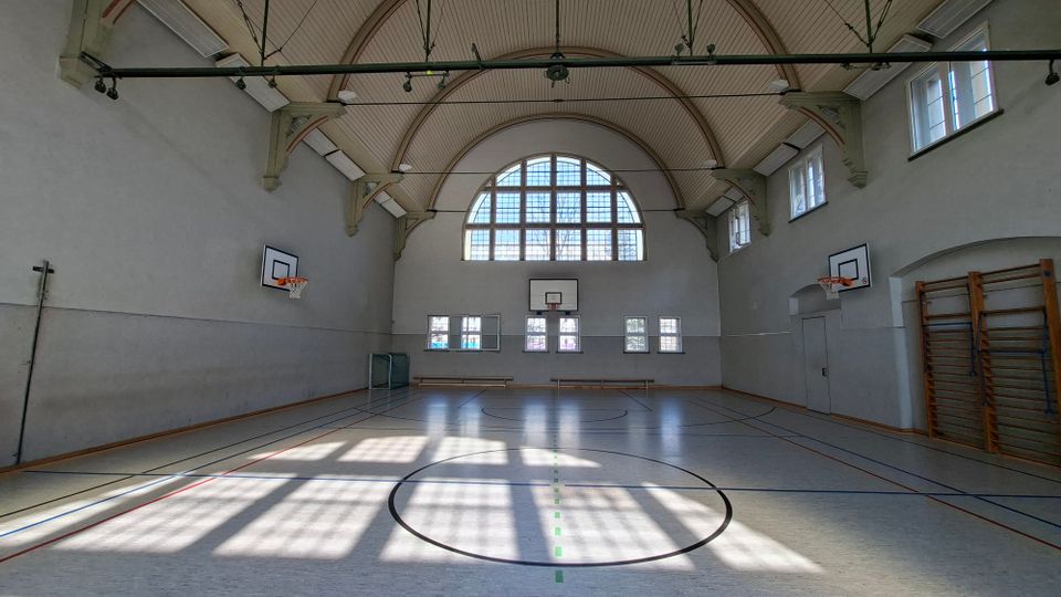 Große Sporthalle mit hoher, gewölbter Decke und großem Bogenfenster an der Stirnseite, durch das viel Tageslicht auf den markierten Hallenboden fällt. An den Wänden sind Basketballkörbe, Turngeräte und mehrere hohe Fenster angebracht.