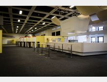 Innenansicht eines großen, modern gestalteten Messe- oder Veranstaltungsraums mit gelben Wänden und hohen Stehtischen mit runden Tischplatten. Die Decke ist mit Gitterstrukturen und rechteckigen Lichtpanelen ausgestattet, während durch große Fenster natürliches Licht einfällt.