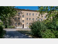 Mehrstöckiges Wohnhaus mit beige gestrichener Fassade und weißen Fensterrahmen, umgeben von Bäumen und Sträuchern. Ein gepflasterter Fußweg führt durch eine grüne Gartenanlage zum Gebäude.