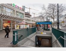 U-Bahn-Eingang Kurfürstendamm in Berlin, umgeben von Einkaufsläden und Passanten. Im Hintergrund moderne Gebäude, Straßenschilder der Linien U1 und U9 sowie Kräne und winterliche Bäume.