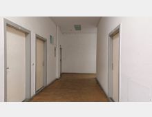 Ein einfacher, heller Flur mit weißen Wänden, einer Decke mit einem rechteckigen Deckenlicht und einem braunen Bodenbelag. Links und rechts befinden sich Türen mit grauen Türrahmen, und im Hintergrund ist eine weitere Tür erkennbar.