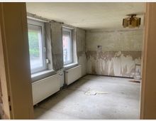 Ein leerer, renovierungsbedürftiger Raum mit zwei großen Fenstern auf der linken Seite, Heizkörpern darunter und abblätterndem Putz an den Wänden. Die Decke ist schlicht, und eine beschädigte Deckenleuchte hängt in der Mitte des Raumes.