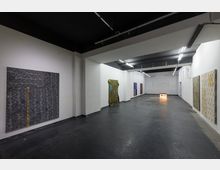 Ein minimalistisch gestalteter Ausstellungsraum mit weißen Wänden, schwarzem Boden und einer dunklen Decke. An den Wänden hängen großformatige, gemusterte Kunstwerke, und im Hintergrund ist ein beleuchtetes Schild zu sehen.