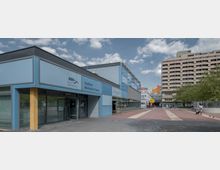 Ein modernes Stadtbad mit hellblauer Fassade und großen Glasfronten im Märkischen Viertel. Im Hintergrund befindet sich ein mehrstöckiges Wohngebäude sowie ein öffentlicher Platz mit Bäumen und Fußgängerwegen.