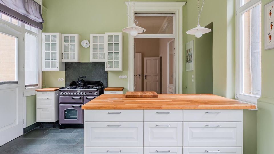 Helle Küche mit salbeigrünen Wänden, hohen Fenstern und weißer Einbauküche mit Glasoberschränken. In der Mitte steht eine große Kücheninsel mit Holzarbeitsplatte; dahinter ein schwarzer Standherd und zwei Pendelleuchten.