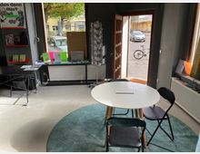 Ein kleiner Raum mit modernem Design, der durch eine offene Holztür auf eine gepflasterte Straße und ein Fahrrad blicken lässt. Im Vordergrund befindet sich ein runder weißer Tisch mit schwarzen Klappstühlen auf einem türkisfarbenen Teppich, links ein Tisch mit Infomaterialien und Regale an der Wand.