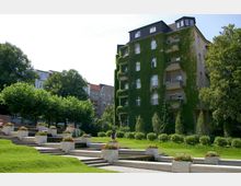 Wohnhaus mit begrünter Fassade und Balkonen, umgeben von Bäumen und Rasenflächen. Im Vordergrund eine terrassenartige Anlage mit Wasserbecken und bepflanzten Blumenkübeln.