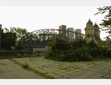 Eine Stahlbogenbrücke mit gemauerten Türmen an den Enden, umgeben von Bäumen und Industriebauten mit großen Silos im Hintergrund. Im Vordergrund ist eine teils bewachsene, gepflasterte Fläche zu sehen.