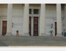 Fassade eines historischen Gebäudes mit großen korinthischen Säulen, breiter Steintreppe und hohen Holztüren. Zwei Blumentöpfe sowie Wandlaternen flankieren den Eingangsbereich.