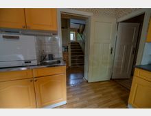 Blick in eine kleine Küche mit gelben Holzschränken, Geschirrspüler und Spüle vor einer gefliesten Rückwand. Durch eine offene Tür ist ein Flur mit Holztreppe und einem Fenster am Ende zu sehen.