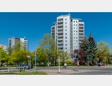 Wohngebiet mit mehrstöckigen, modernen Wohnhochhäusern, umgeben von Bäumen und einer Straße im Vordergrund mit Ampeln und mehreren parkenden Autos. Ein Radfahrer und Spaziergänger befinden sich nahe einer Kreuzung, während der Himmel klar und blau ist.