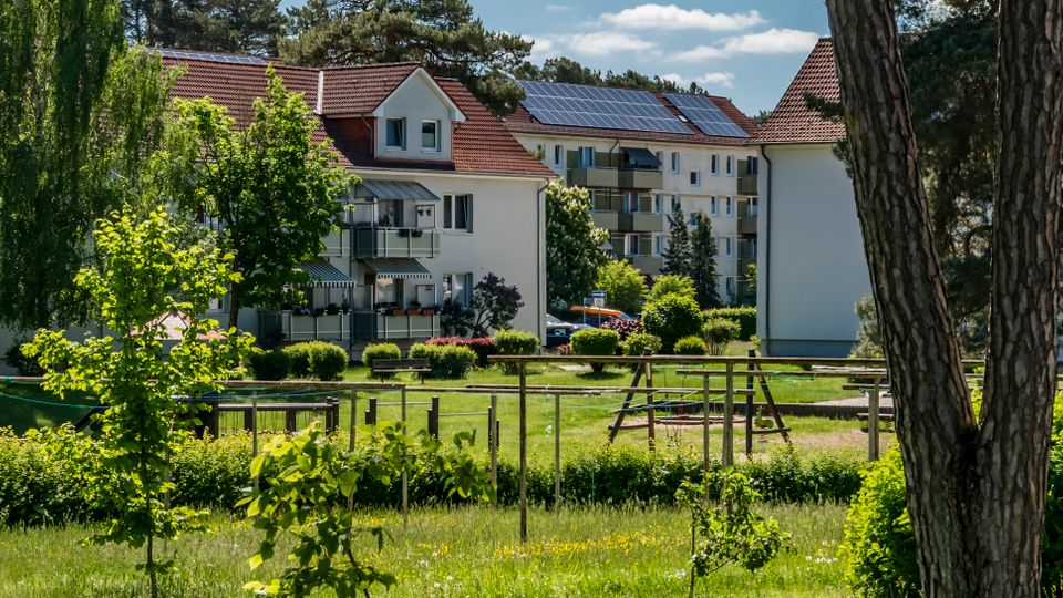 Wohnanlage mit mehrstöckigen Wohnhäusern mit roten Ziegeldächern, Balkonen und teils Solarpaneelen auf den Dächern. Davor liegt eine grüne Parkfläche mit Bäumen, Hecken und einem Spielplatzbereich mit Klettergerüst und Wegen.