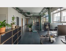 Moderner Büroarbeitsbereich mit Schreibtischen, mehreren Computerbildschirmen und dekorativen grünen Pflanzen. Im Hintergrund befinden sich schallisolierte Kabinen, große Fenster mit Blick auf ein Nachbargebäude und eine sichtbare Deckeninstallation mit Kabelkanälen.