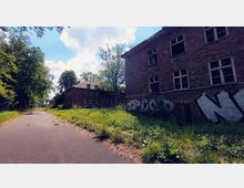 Eine verlassene Backsteinanlage mit mehreren Gebäuden, deren Fenster teils beschädigt oder offen sind. Die Umgebung ist von Gras, wilder Vegetation und einer asphaltierten Straße geprägt; eine Fassade ist mit Graffiti bedeckt, und Bäume rahmen das Gelände ein.